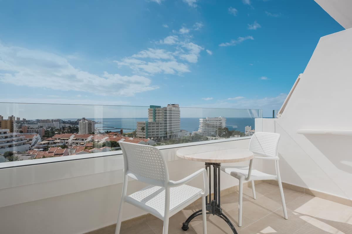 A balcony with two white chairs and a round table, overlooking a coastal cityscape with buildings and the sea.