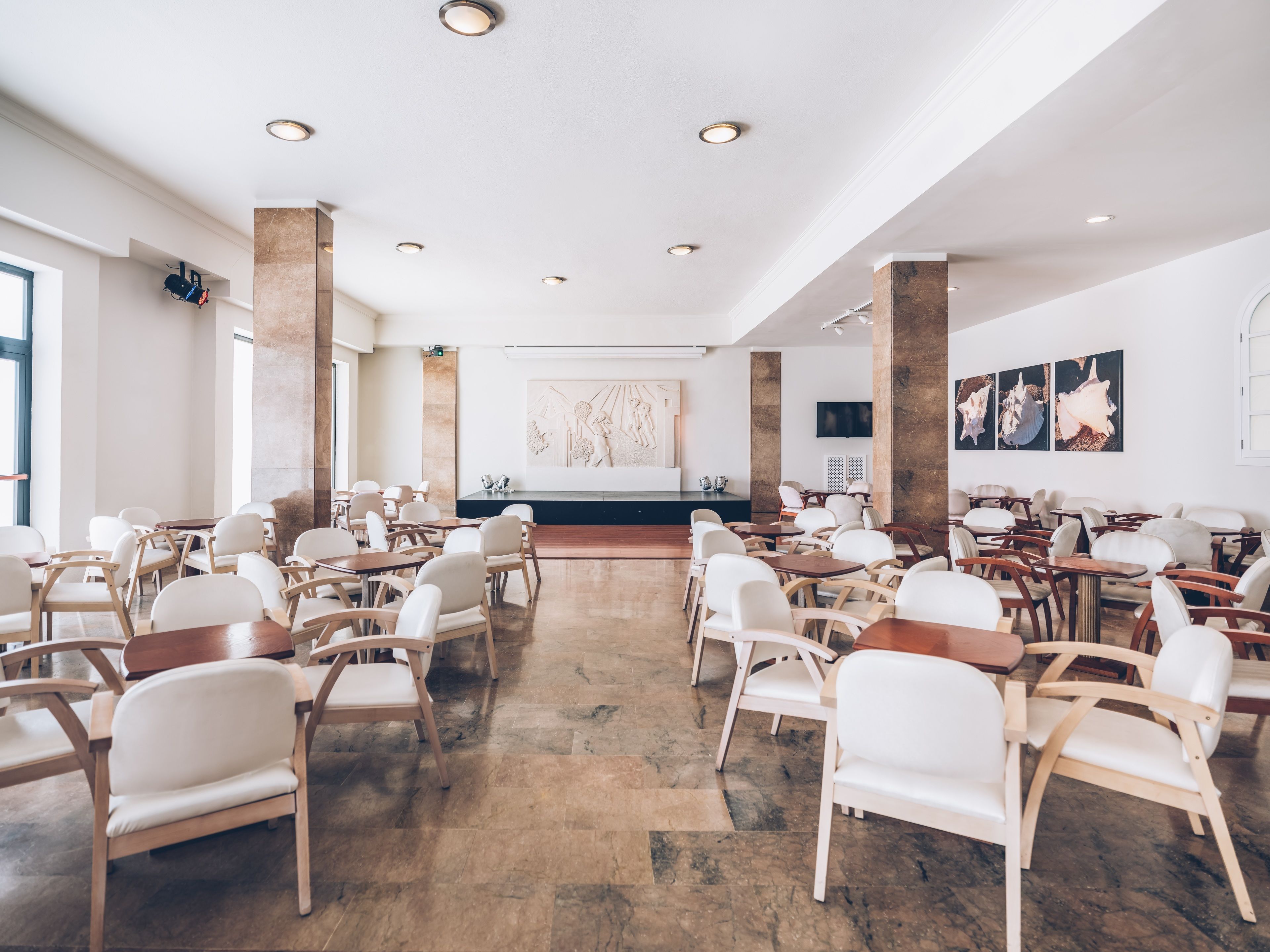 A spacious conference room with white chairs and tables, a stage with a podium, and a large wall art piece.