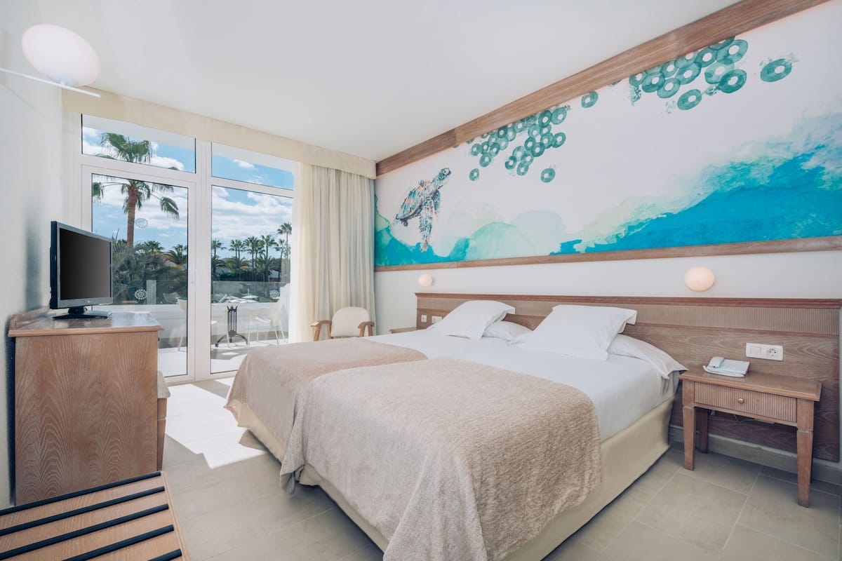 A hotel room with two beds, a TV, a desk, and a large window with a view of palm trees.
