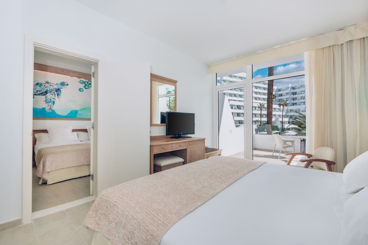 A hotel room with a bed, a desk with a television, a chair, and a balcony with a view of a building.