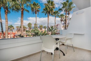 A balcony with two white wicker chairs and a small round table, overlooking a cityscape with palm trees and buildings.
