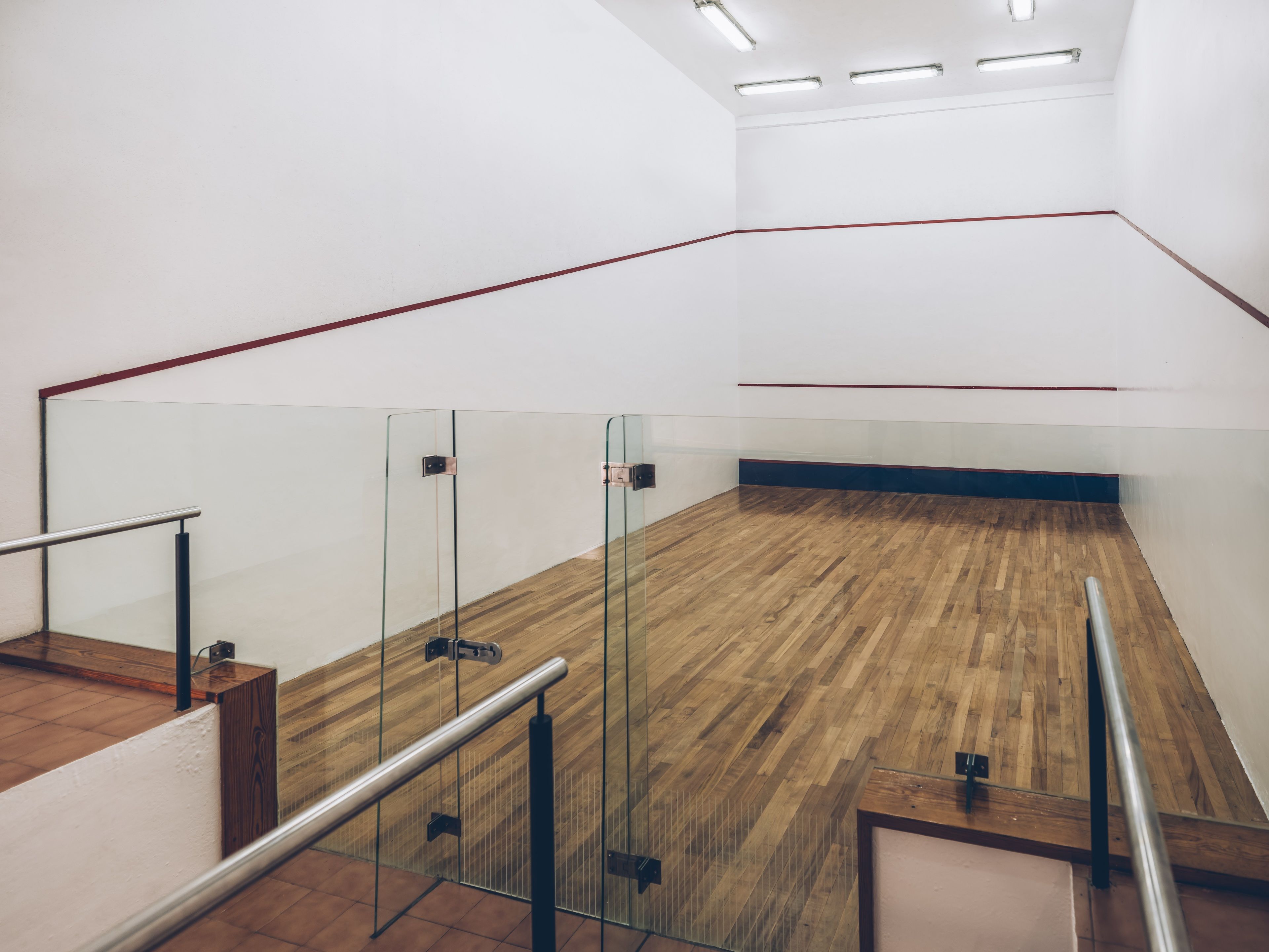 Una cancha de squash con paredes de vidrio, suelo de madera y barandillas metálicas.