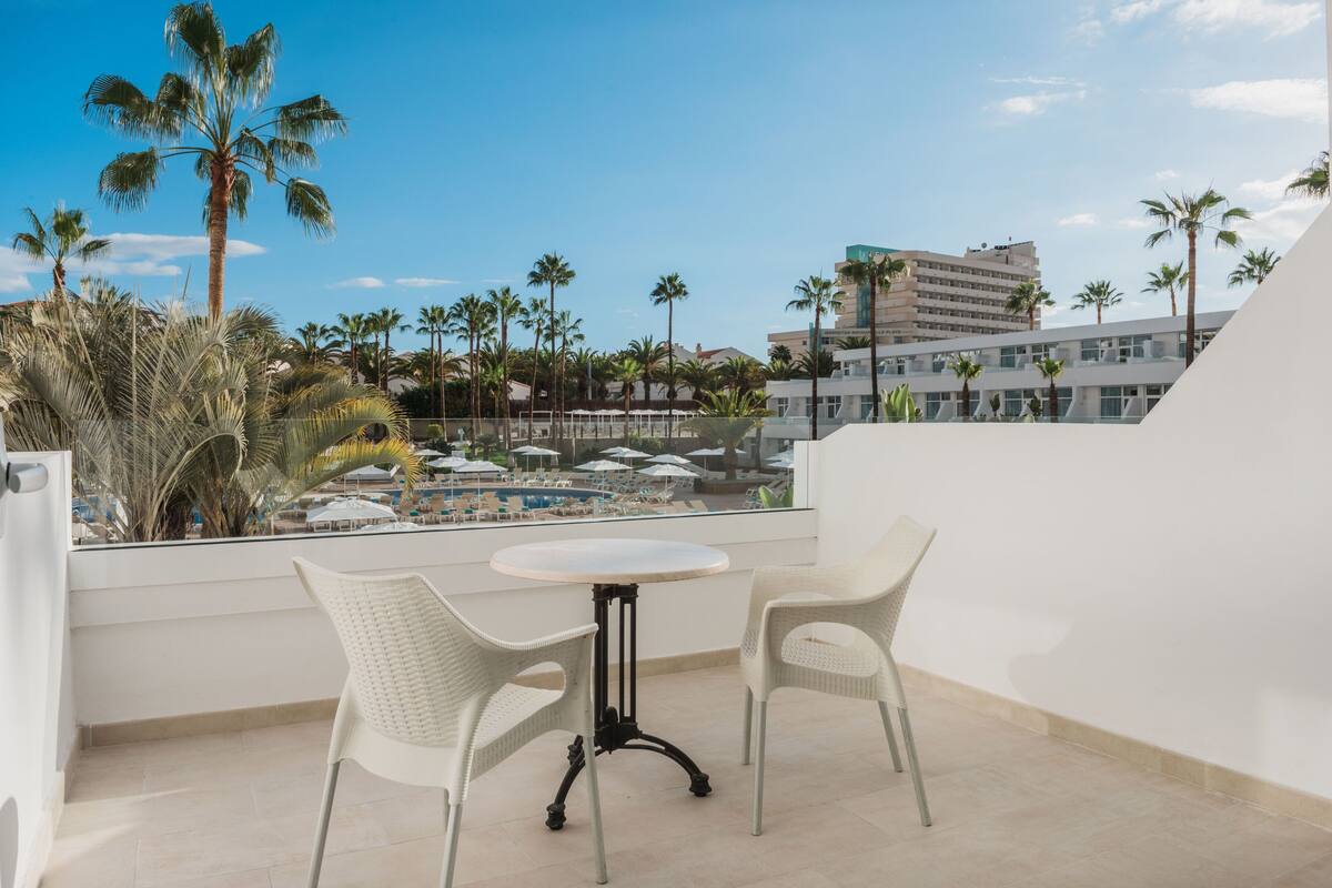 A balcony with two white wicker chairs and a small round table, overlooking a pool area with umbrellas and palm trees.