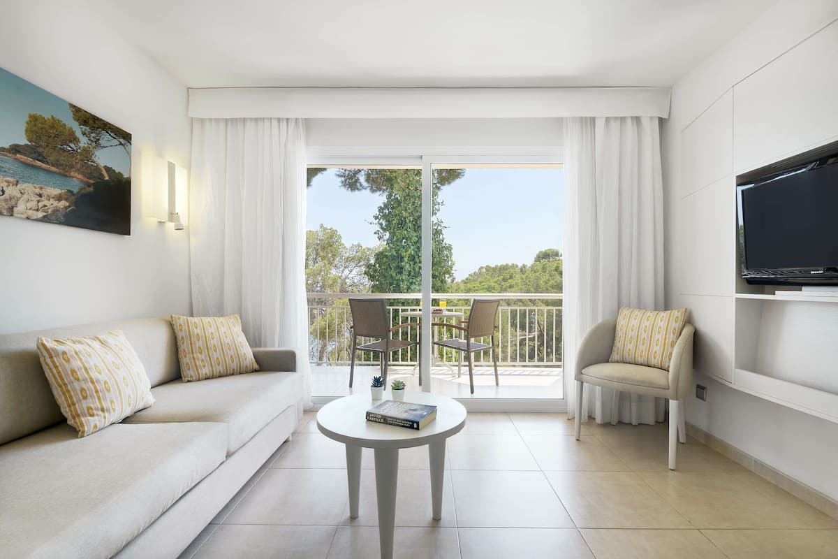 A modern living room with a sofa, a small round table, and a chair. There is a balcony with a view of trees and a body of water.