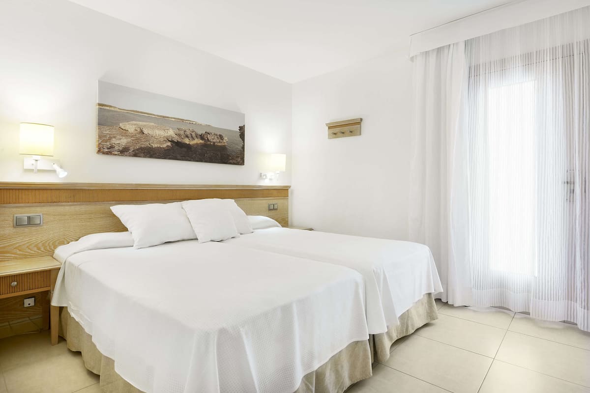 A hotel room with two beds, a wooden headboard, a painting on the wall, and a window with sheer curtains.