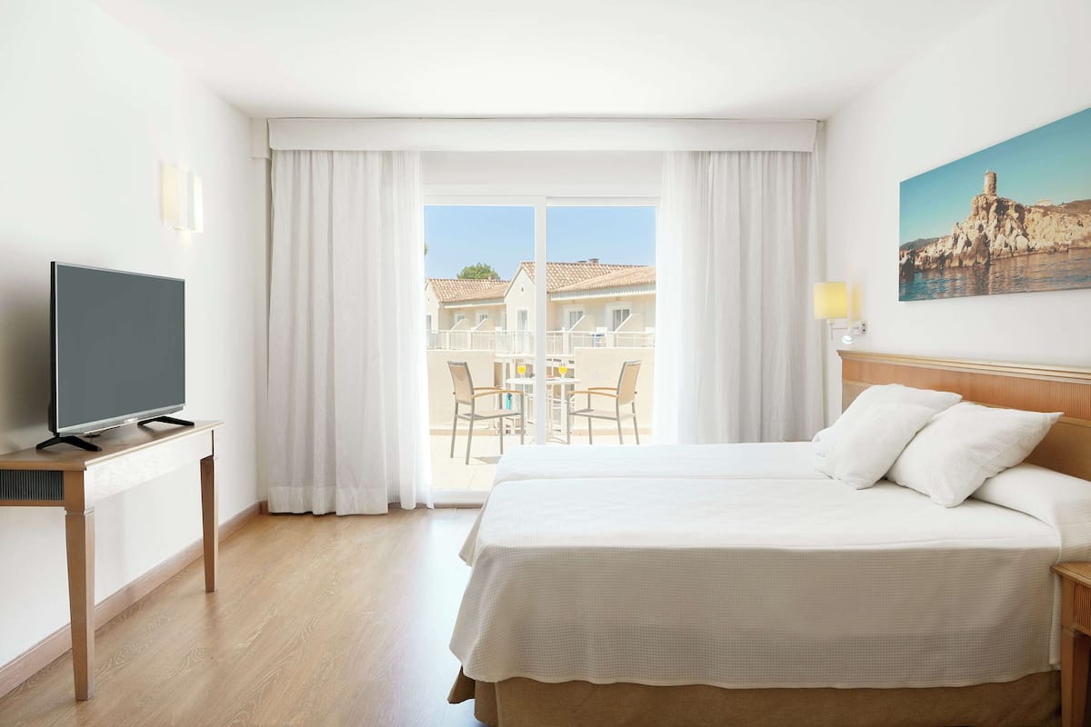 A hotel room with a bed, a TV on a stand, a balcony with chairs, and a painting on the wall.