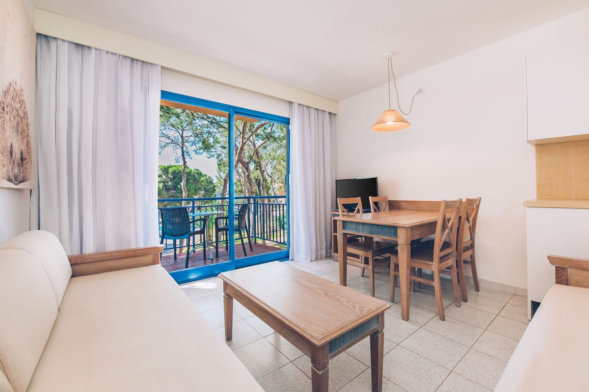 A modern room with a sofa, a wooden dining table with chairs, a TV, and a balcony with a view of trees.