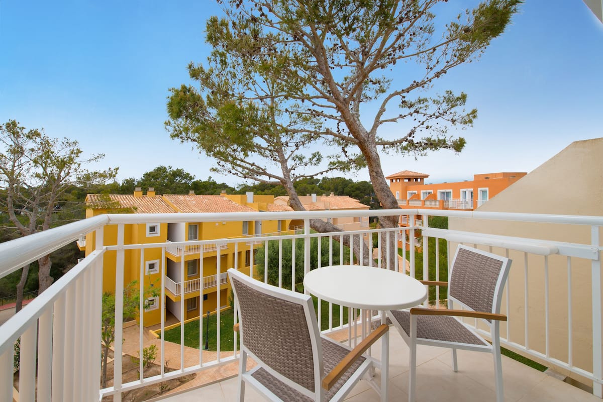 A balcony with wicker chairs and a small table, overlooking a residential area with yellow buildings and a tree.