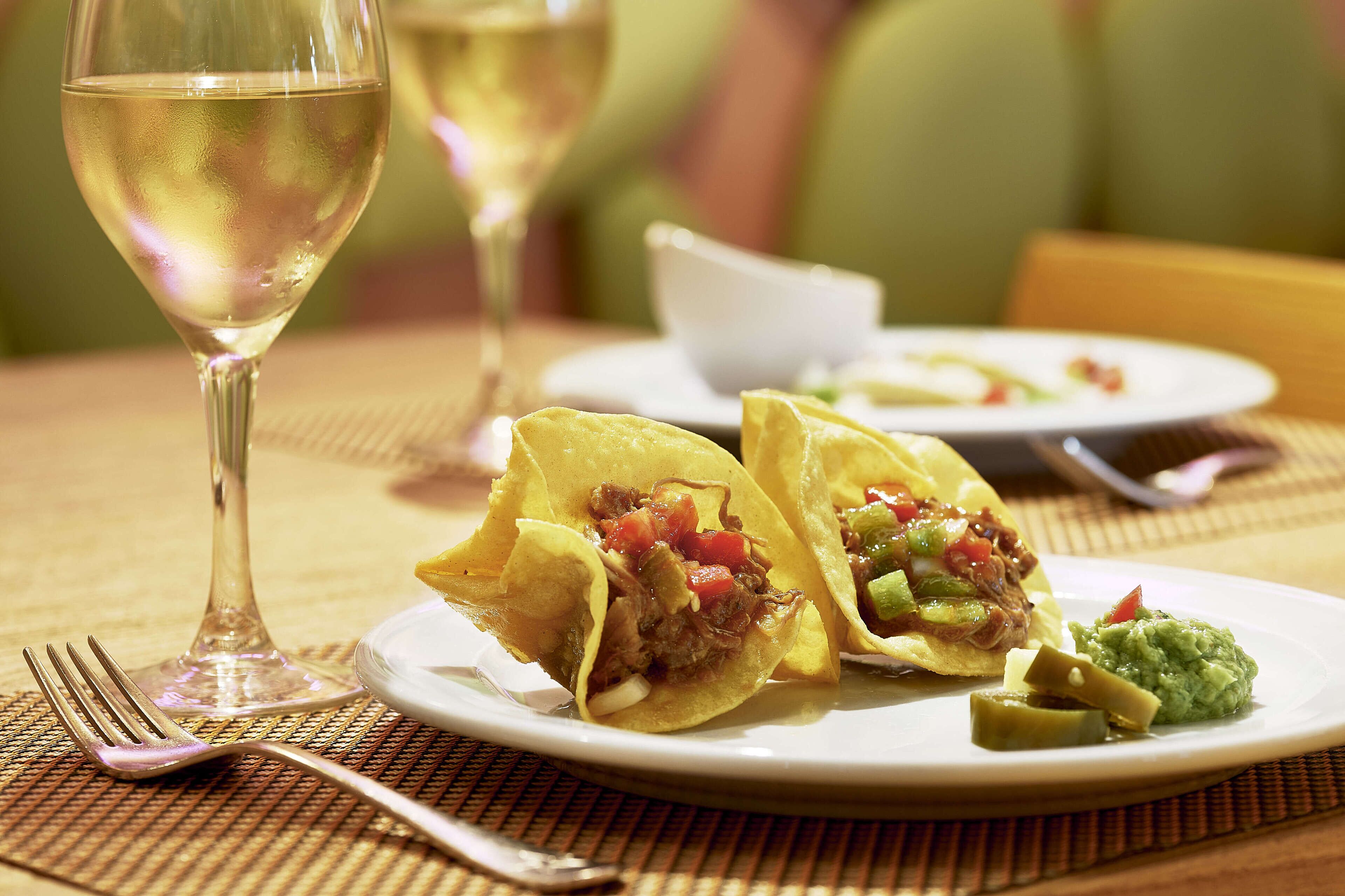 Una mesa de comedor con un plato de tacos, una copa de vino blanco y un tenedor.