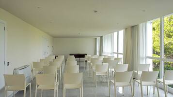 Un salón de conferencias con filas de sillas blancas, una mesa negra y grandes ventanas con cortinas.