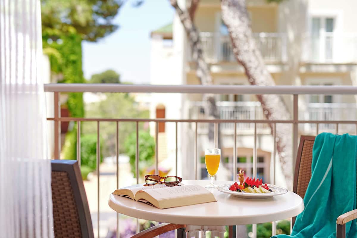 A round table with a book, a glass of orange juice, and a plate of fruit on a balcony with a view of buildings and trees.
