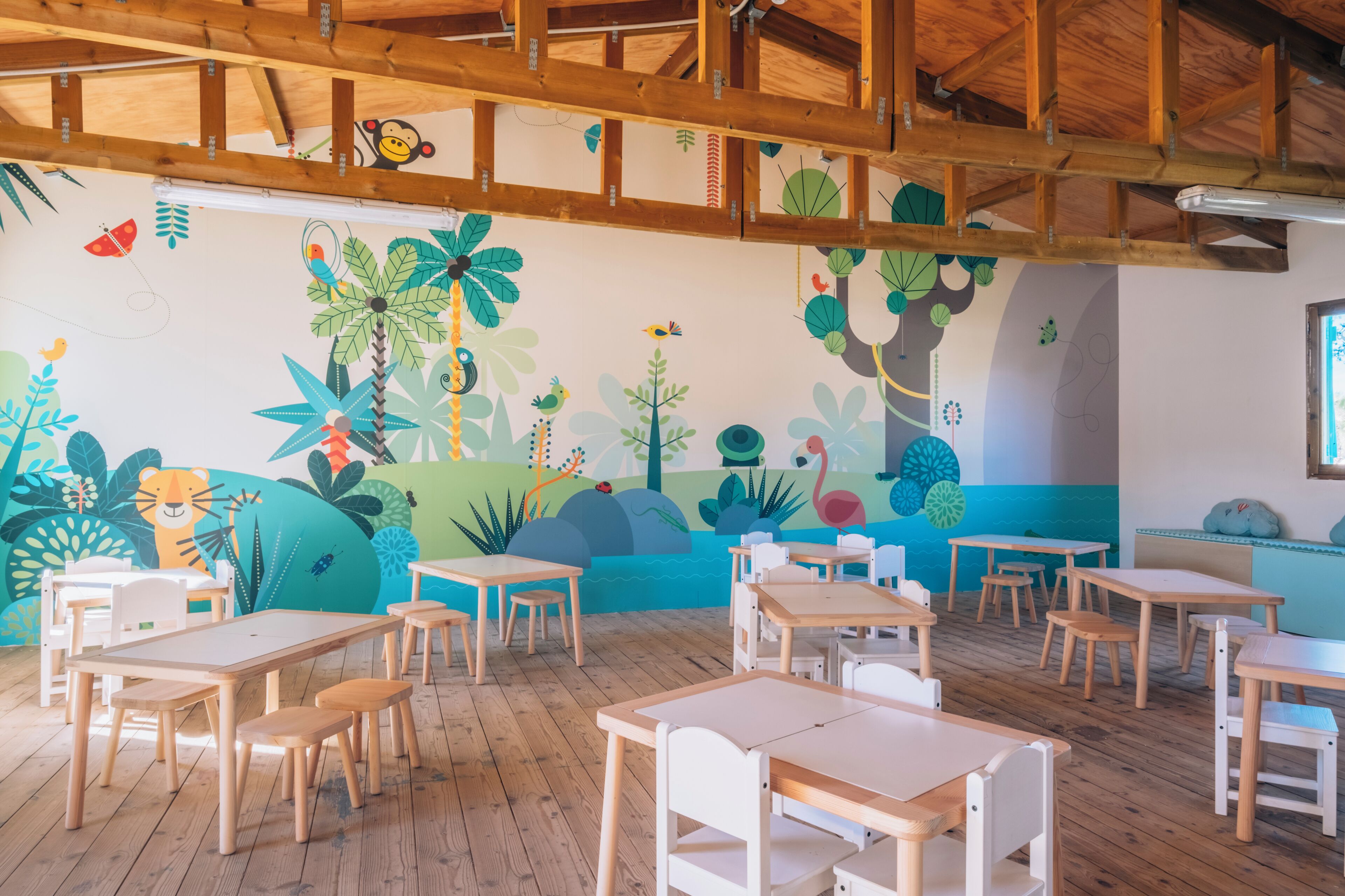 A classroom with wooden tables and chairs, a mural with tropical and animal designs, and a wooden ceiling structure.