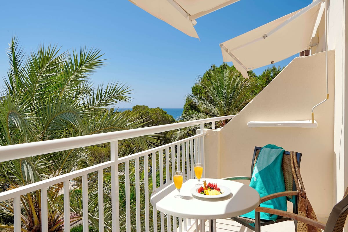 A balcony with a white railing, a small table with a plate of food and two glasses of orange juice, and a chair with a blue towel.