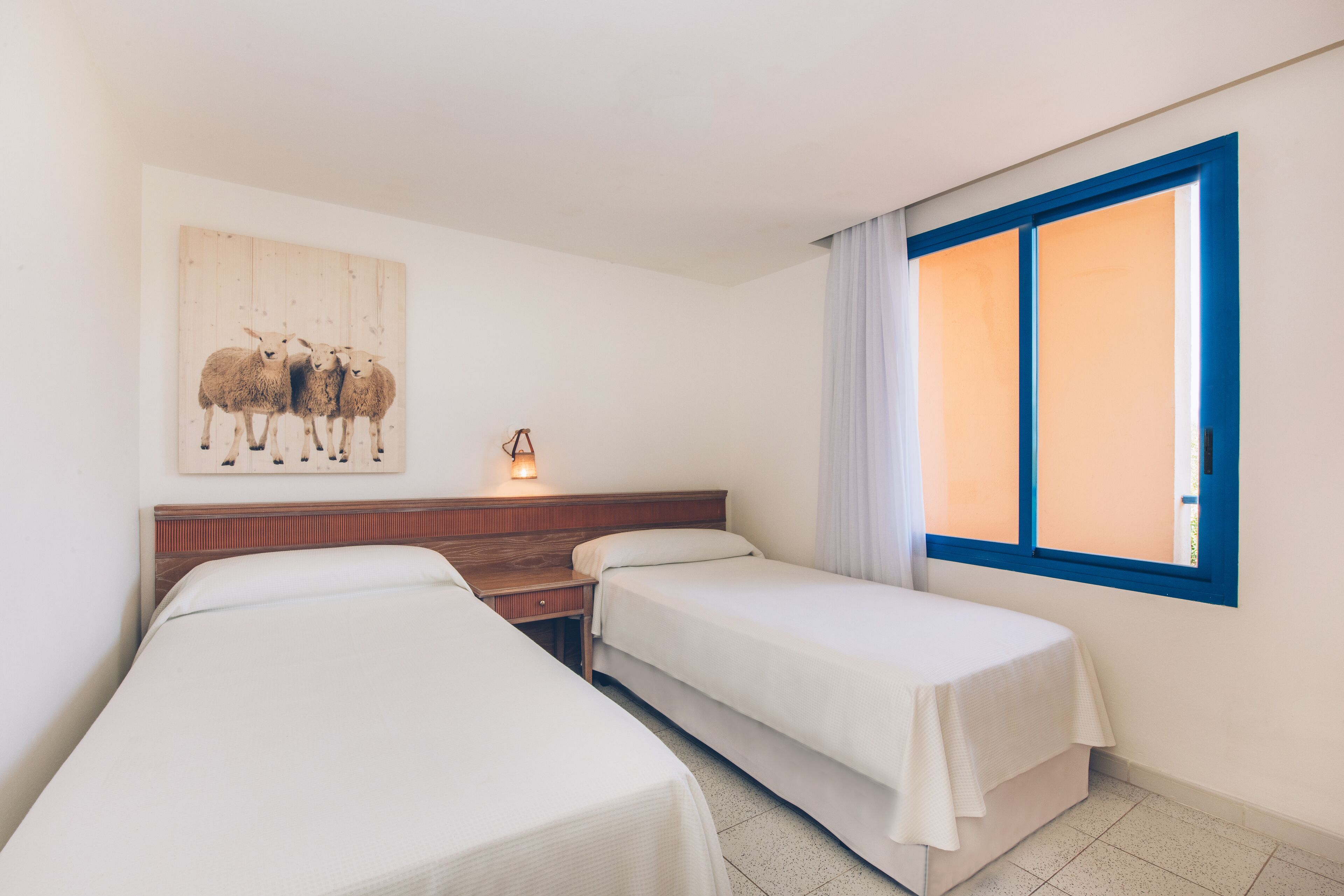 Een hotelkamer met twee eenpersoonsbedden, een houten hoofdbord, een schilderij van schapen aan de muur, en een raam met blauwe rand.
