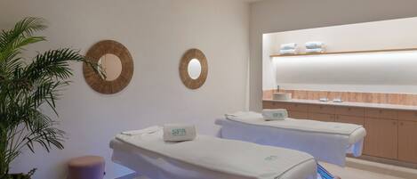A spa room with two treatment tables, a large mirror, and a shelf with towels.