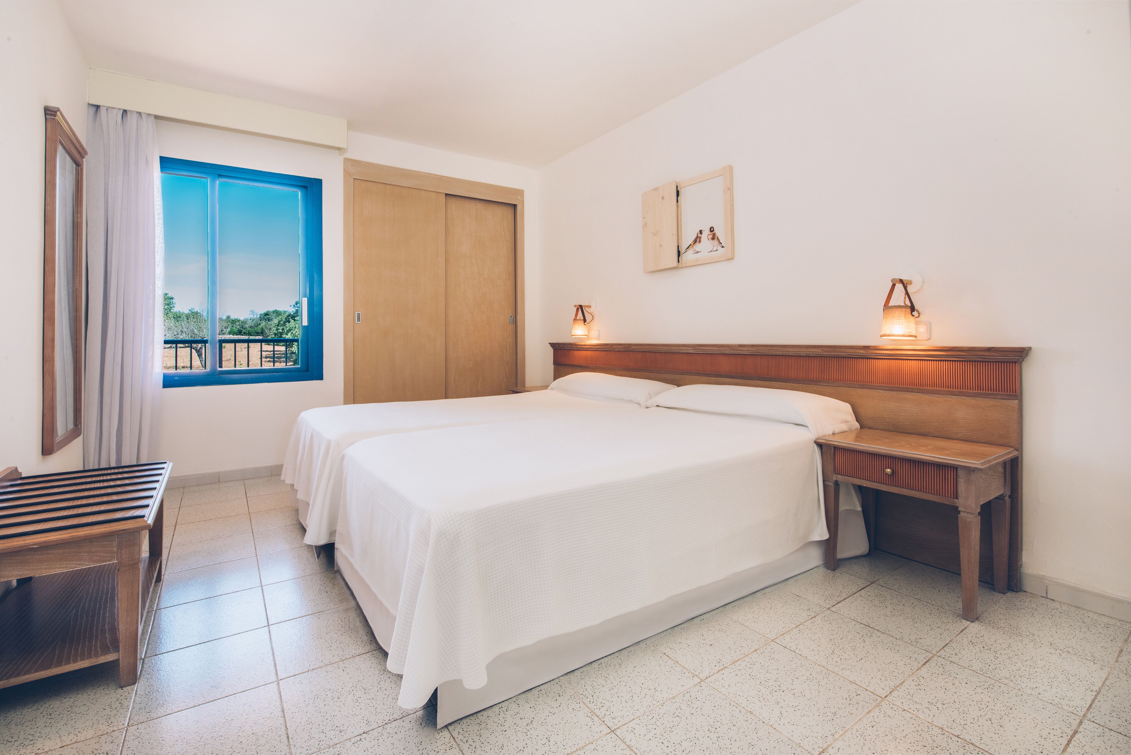 Un dormitorio con una cama grande, una mesita de noche de madera, un banco, una ventana con cortinas y vistas al exterior.