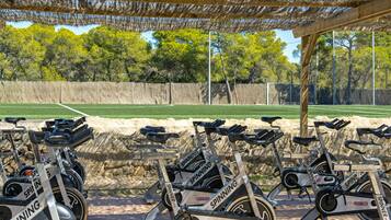 Varias bicicletas estáticas alineadas bajo una pérgola, con una cancha de tenis al fondo.