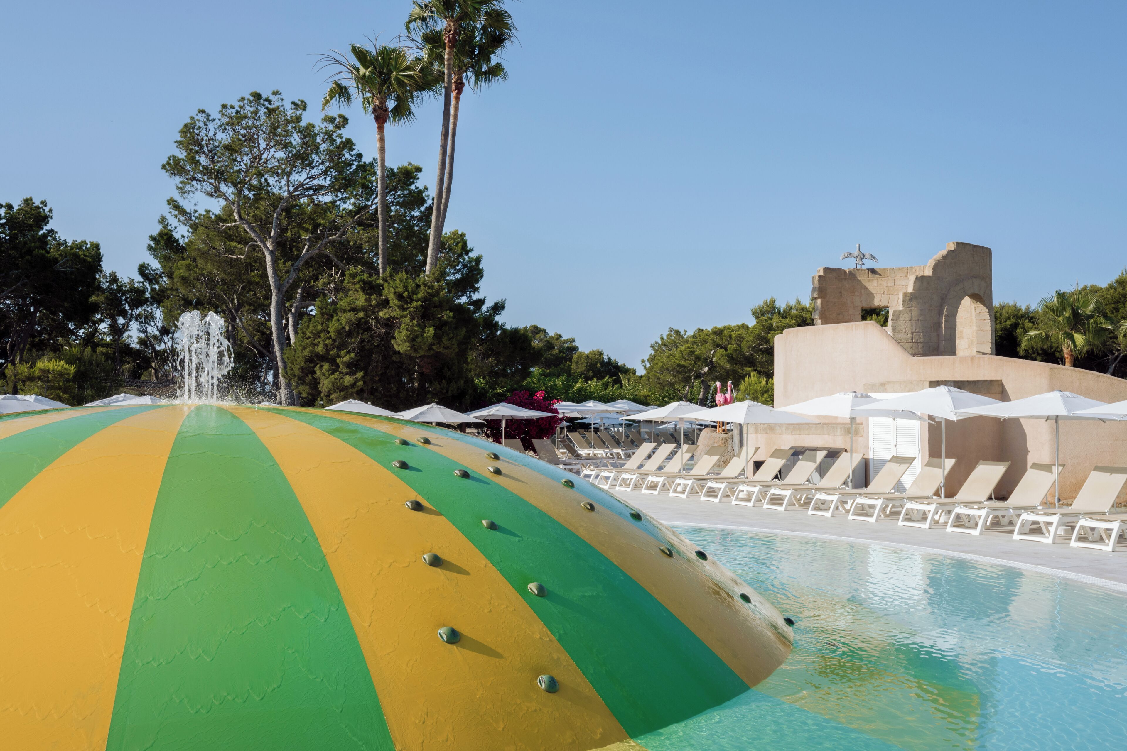 A pool area with a large, colorful, semi-circular structure, lounge chairs, umbrellas, and a fountain.