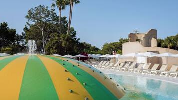 A pool area with a large, colorful, semi-circular structure, lounge chairs, umbrellas, and a fountain.