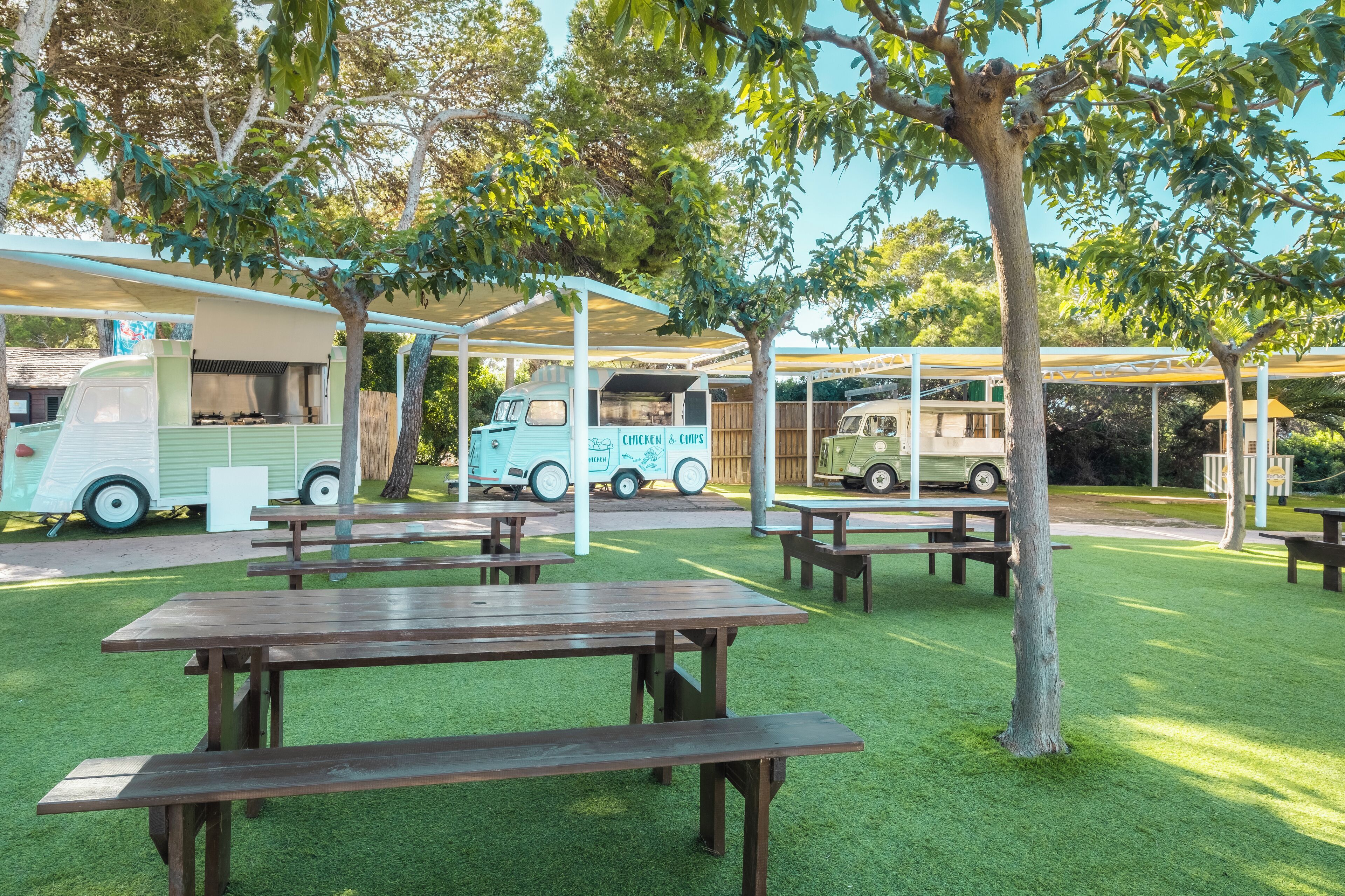 Zona de pícnic amb taules i seients coberts, envoltada d'arbres i un food truck.
