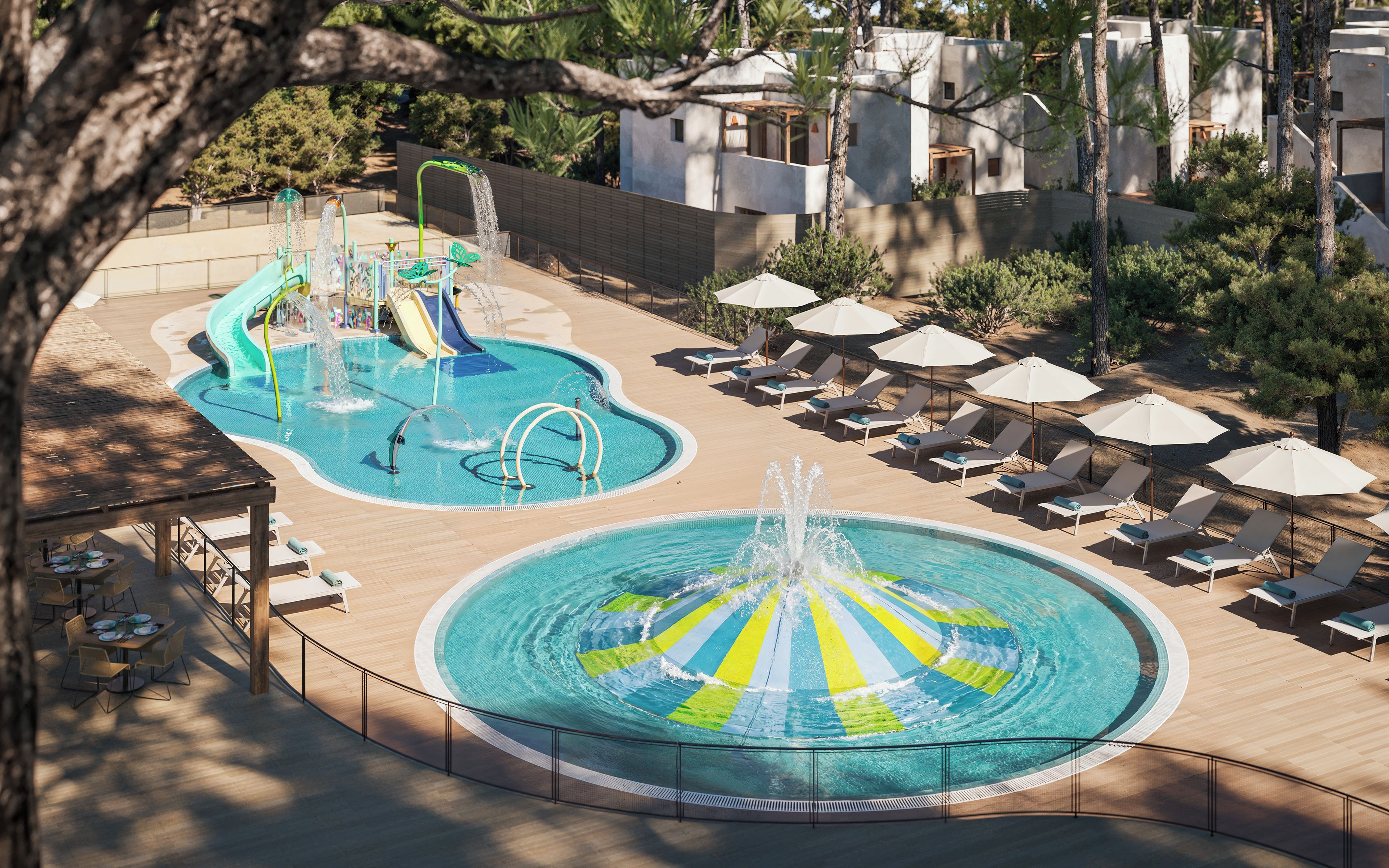 Un área de piscina con varias fuentes de agua, incluyendo una fuente central y un tobogán, rodeada de sillones de descanso y sombrillas.