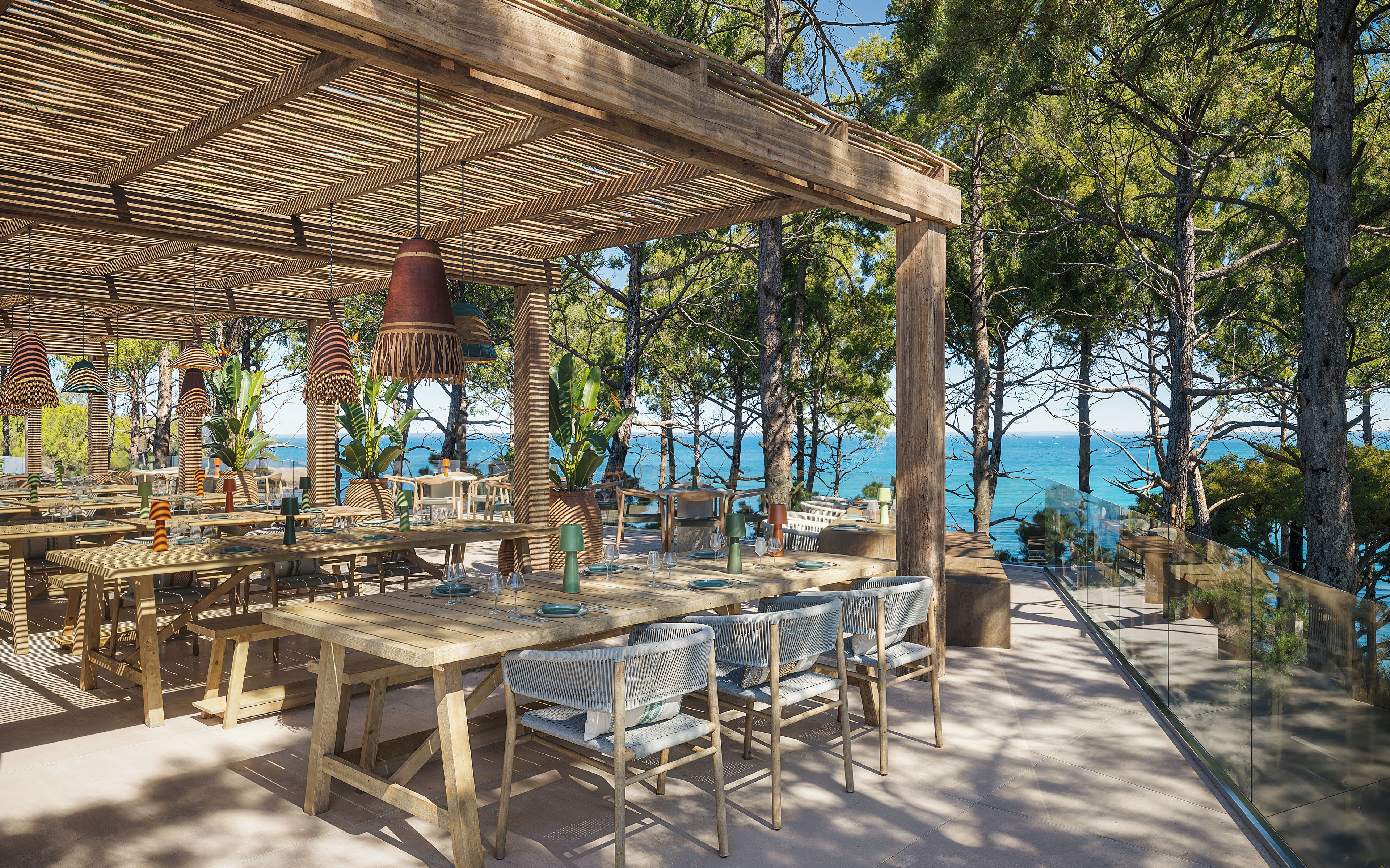 An outdoor dining area with wooden tables and chairs, surrounded by trees and overlooking a body of water.
