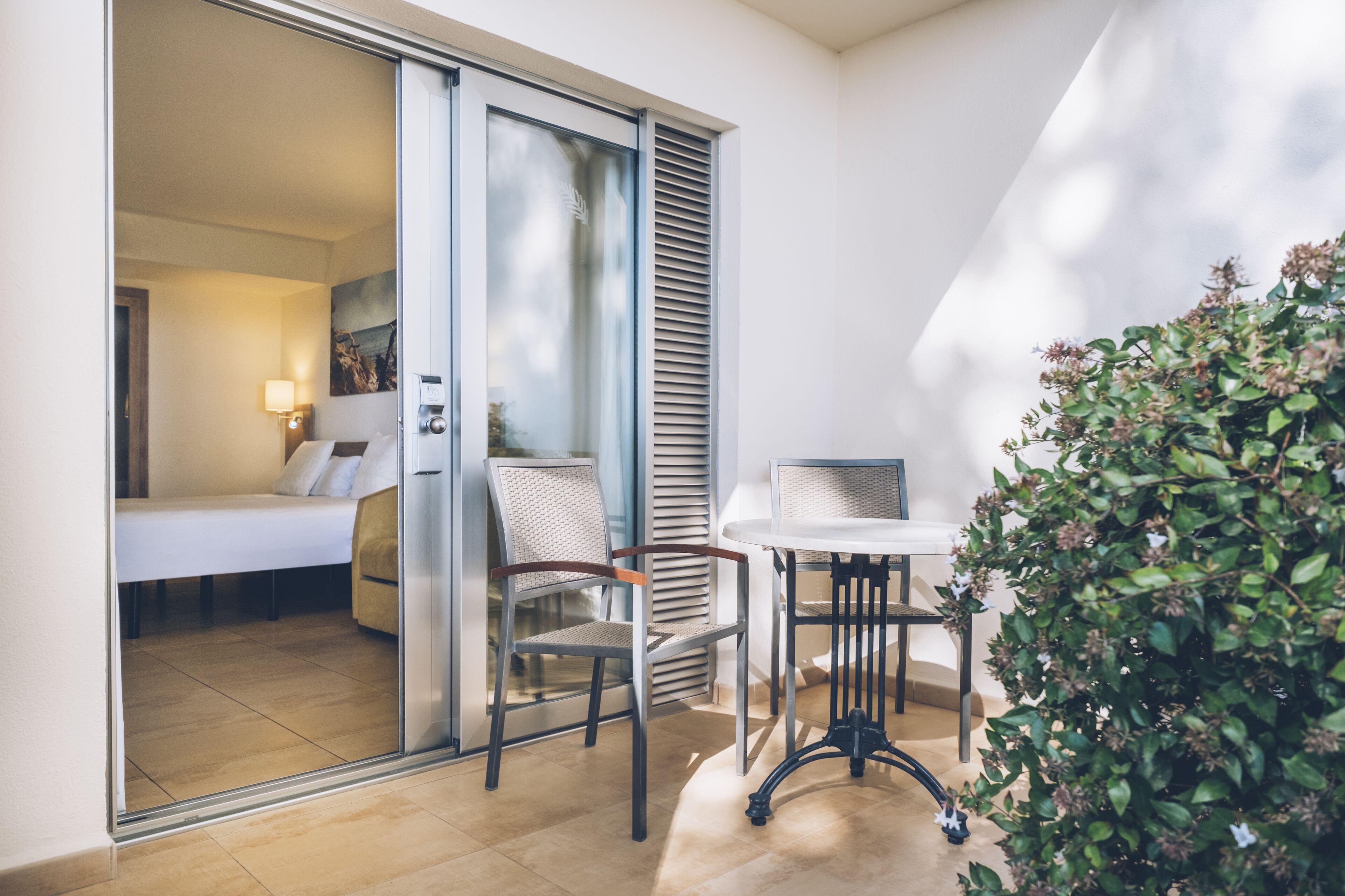 Una habitación de hotel moderna con puerta corrediza de vidrio que da a un balcón, una pequeña mesa con una silla y una planta en maceta.