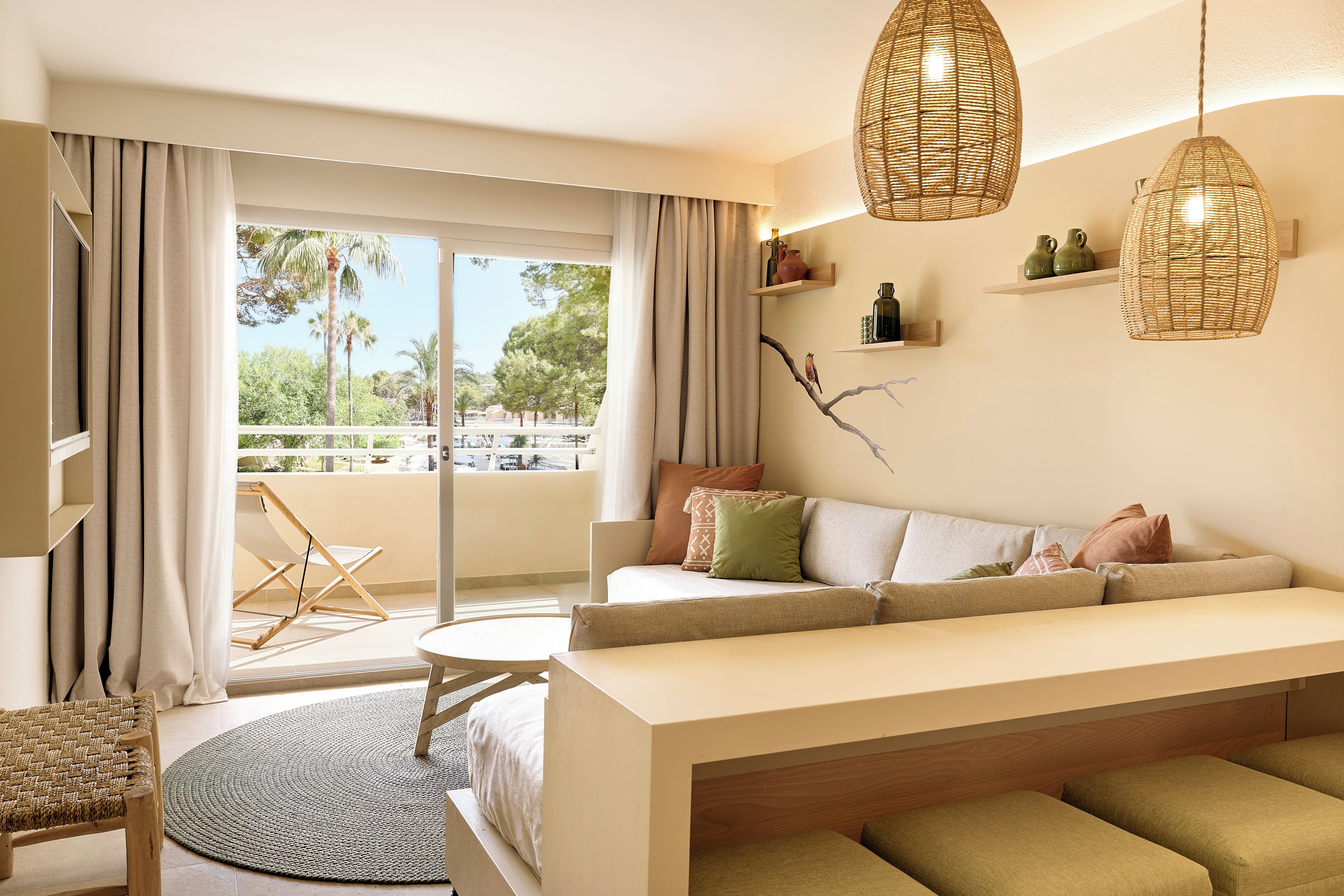 Un salon moderne avec un canapé, une petite table ronde et un fauteuil en osier. On y trouve des suspensions en osier, une vue sur l’extérieur grâce à une grande fenêtre et un balcon avec un fauteuil.