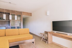 Un salon moderne avec un canapé en bois, un téléviseur à écran plat posé sur un meuble en bois, et un coin cuisine doté de meubles en bois et de luminaires suspendus.