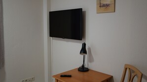 A room with a television mounted on the wall, a desk with a lamp, a remote control, and a chair. - Hotel Milano (Hildesheim)
