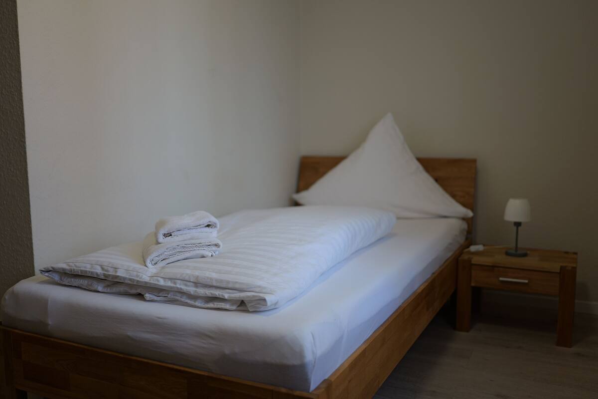 A neatly made bed with white linens and a folded towel, a wooden headboard, a bedside table with a lamp, and a small drawer.