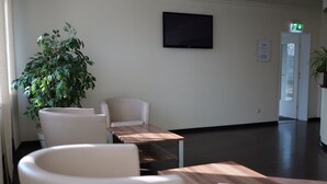 A modern waiting area with a large potted plant, a flat-screen TV mounted on the wall, and a sign indicating the location of rooms. - Hotel Milano (Hildesheim)
