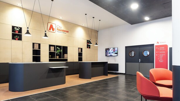 A hotel reception area with a modern design, featuring a reception desk, seating area, and a digital screen displaying information. - City Residence Ivry (Ivry-sur-Seine)