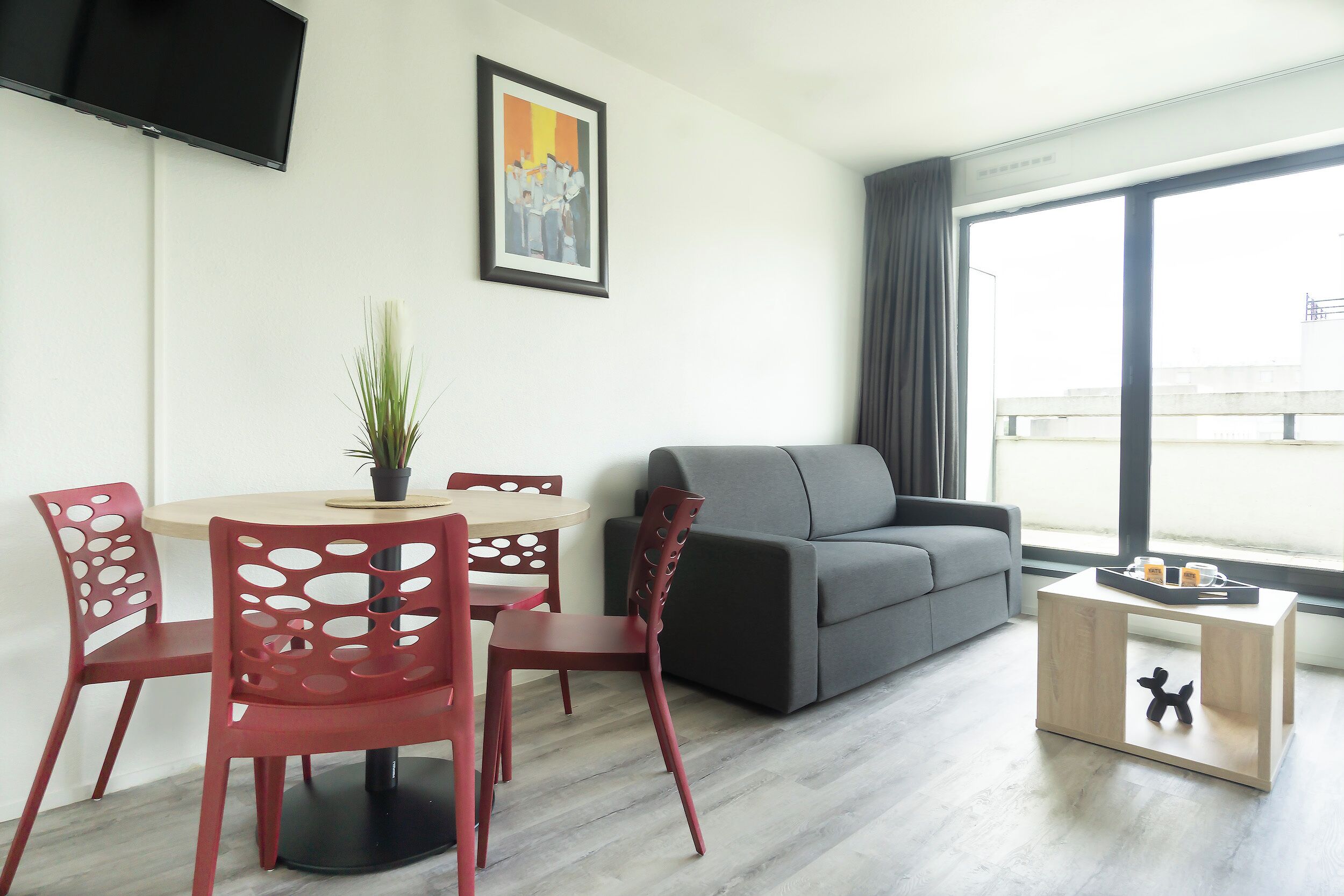 A modern living room with a grey sofa, red chairs, a small round table, and a TV mounted on the wall.