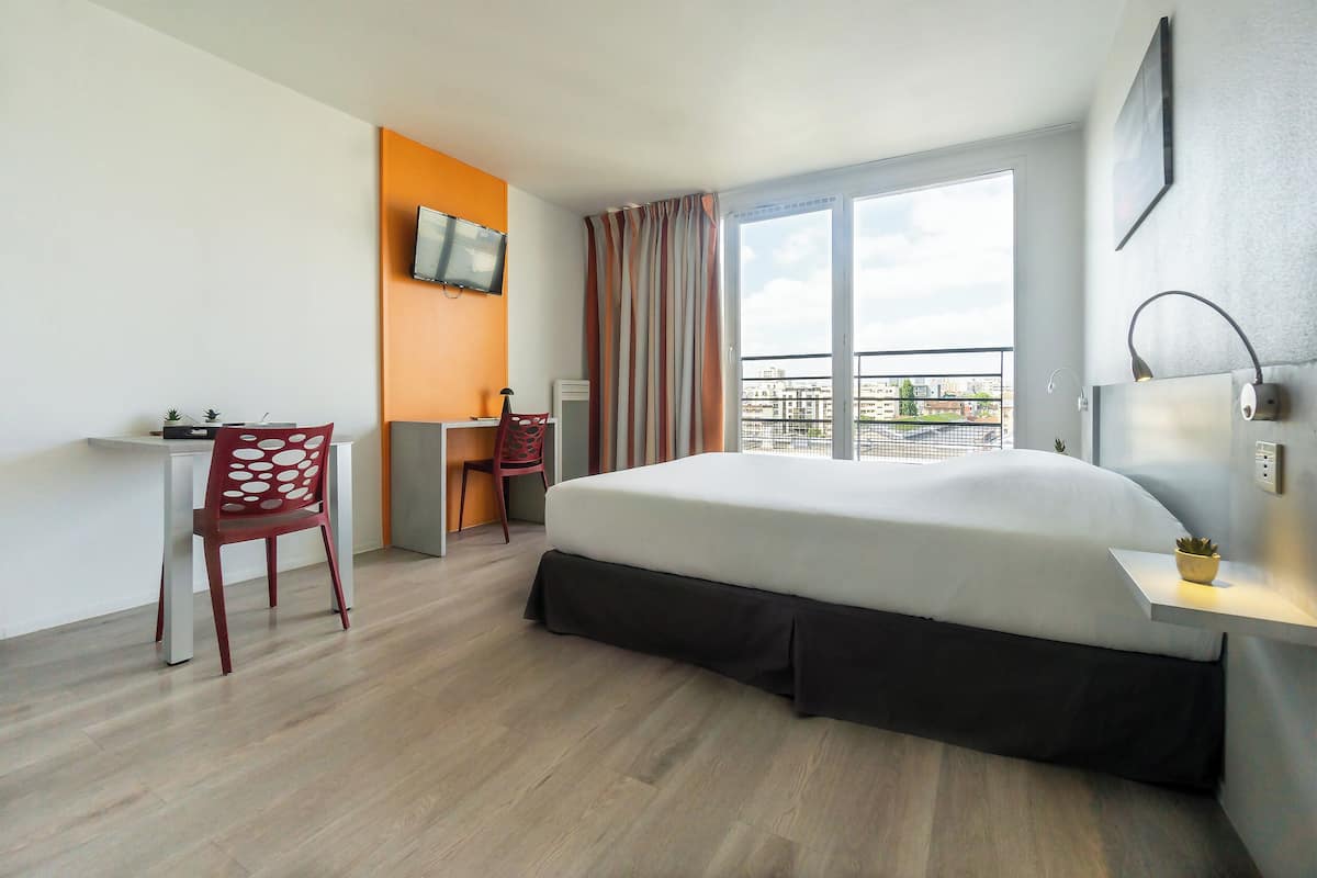 A hotel room with a bed, a desk, a chair, a TV, and a window with a view of buildings.
