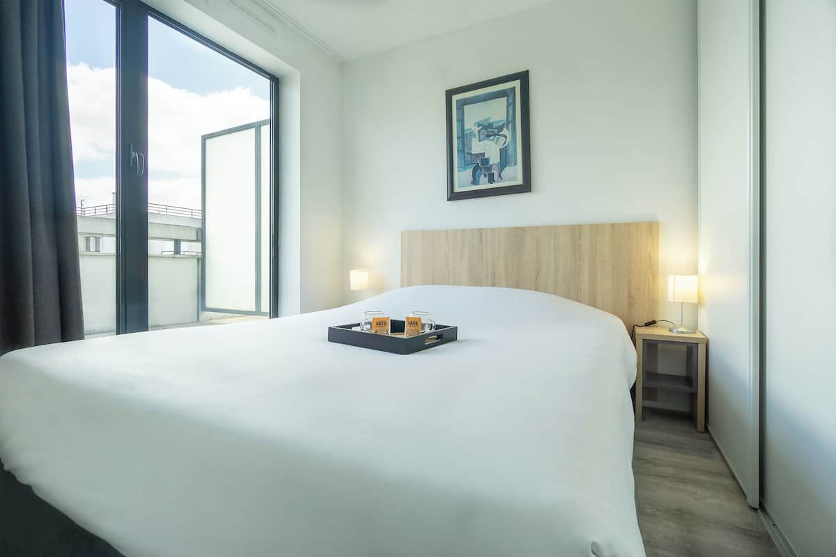 A hotel room with a large bed, a bedside table with a lamp, a tray with two cups, and a framed picture on the wall.