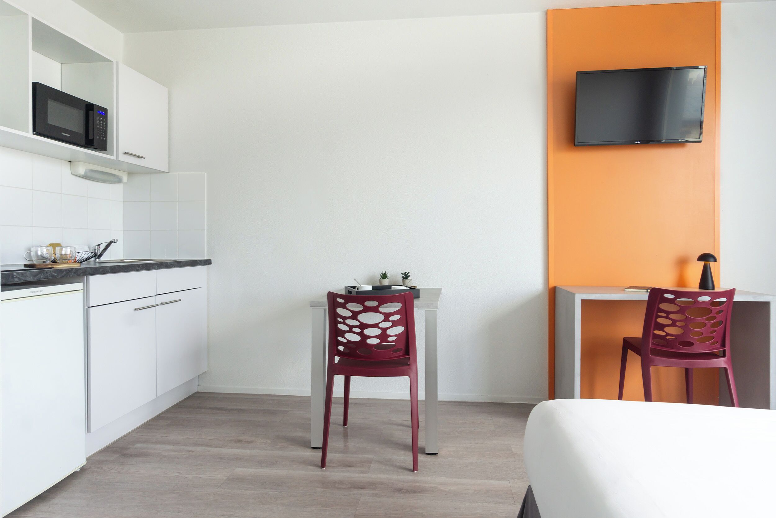Une pièce de taille modeste comprenant un coin cuisine, un lit, deux chaises, un bureau et un téléviseur fixé au mur.
