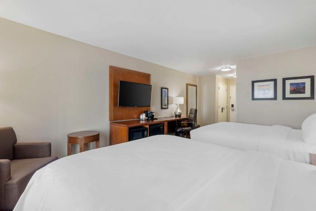 A hotel room with two beds, a desk with a flat-screen TV, a chair, a lamp, and framed pictures on the wall.