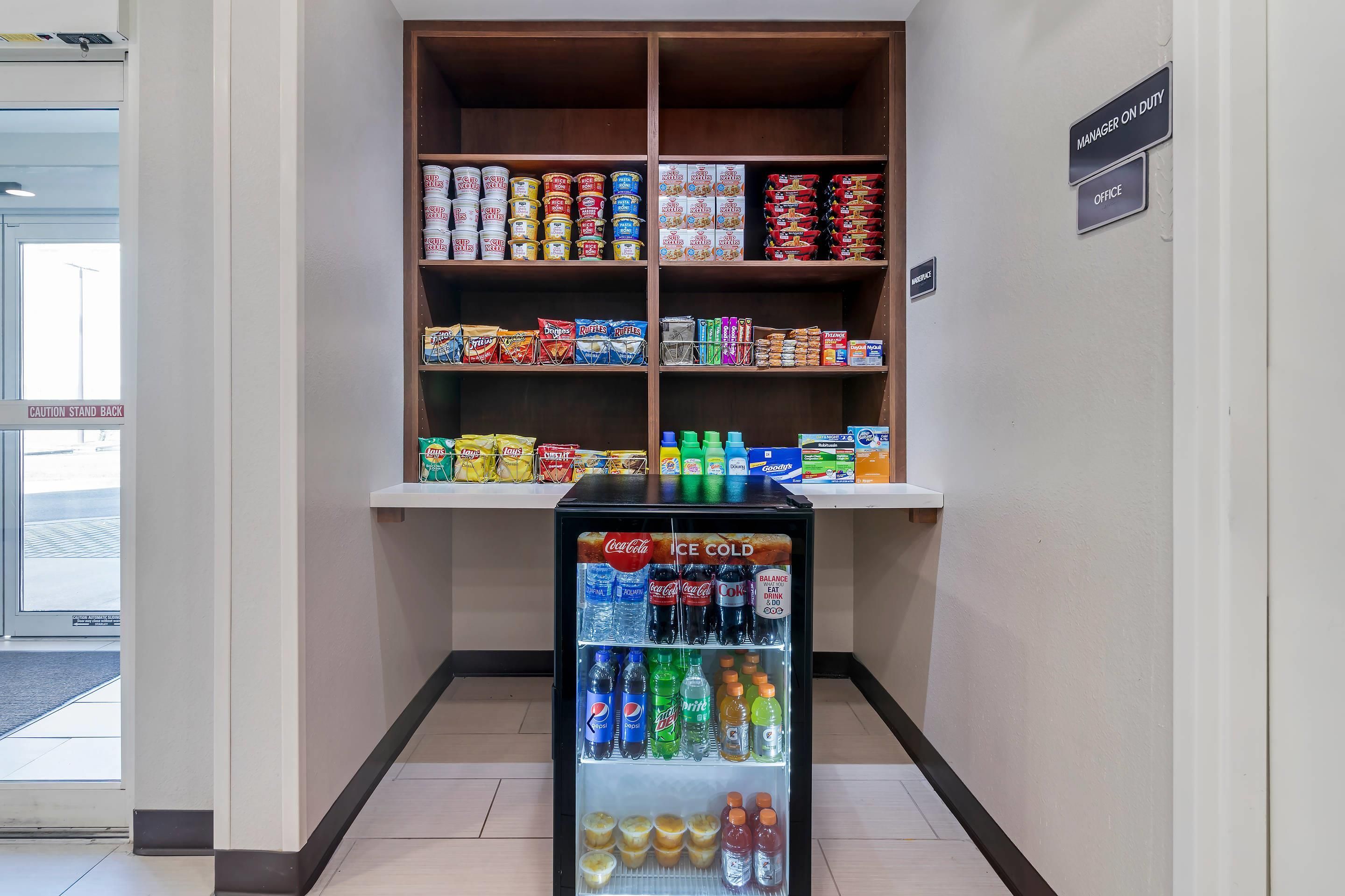 A vending machine stocked with beverages and snacks, located in a hallway with shelves above containing various food items.
