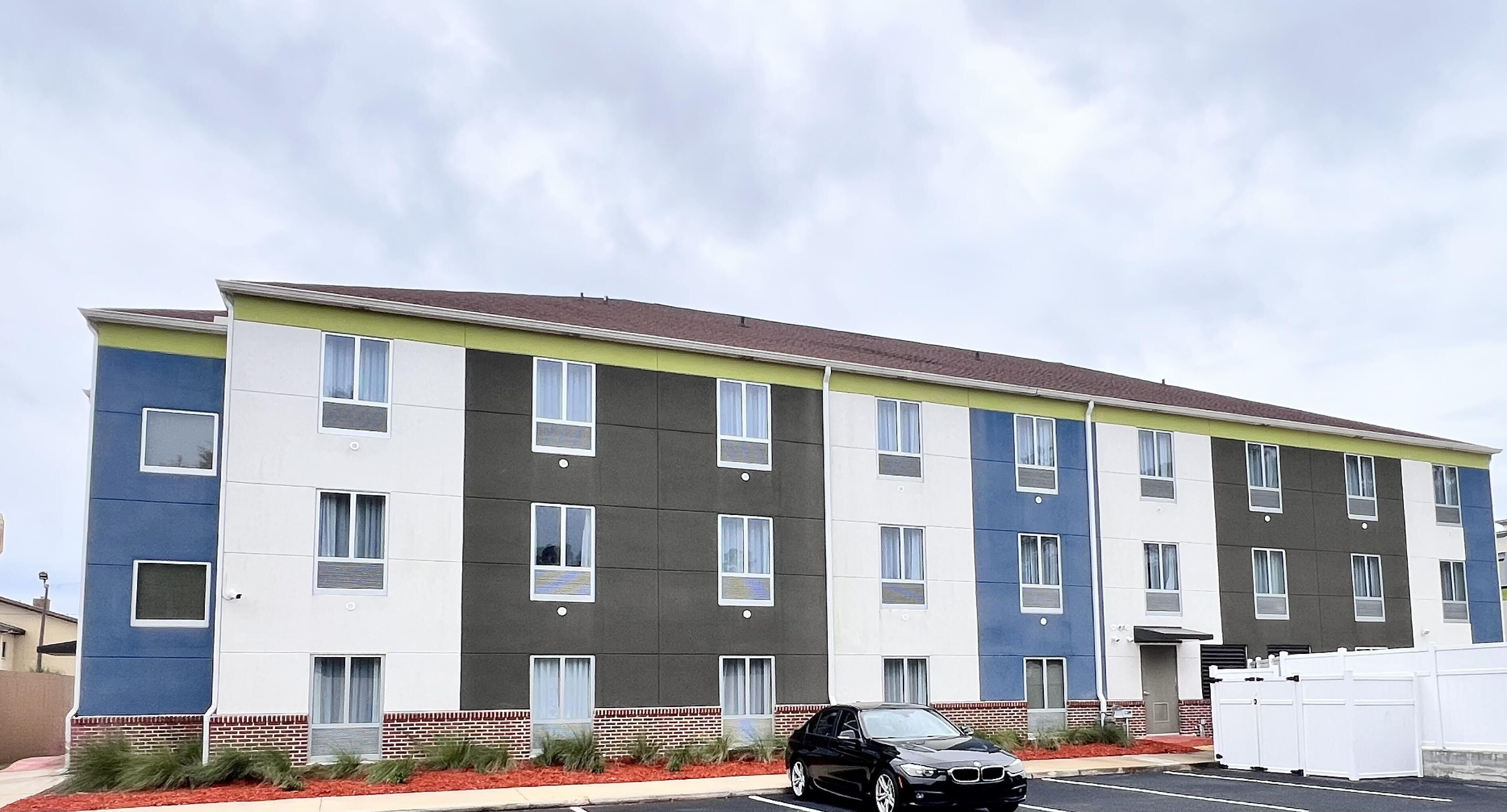 A multi-story building with a modern design, featuring a mix of white, grey, and blue exterior panels, and a flat roof.