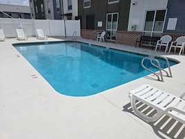 Una piscina rectangular con una escalera de metal, sillas reclinables blancas y un muro de ladrillo.