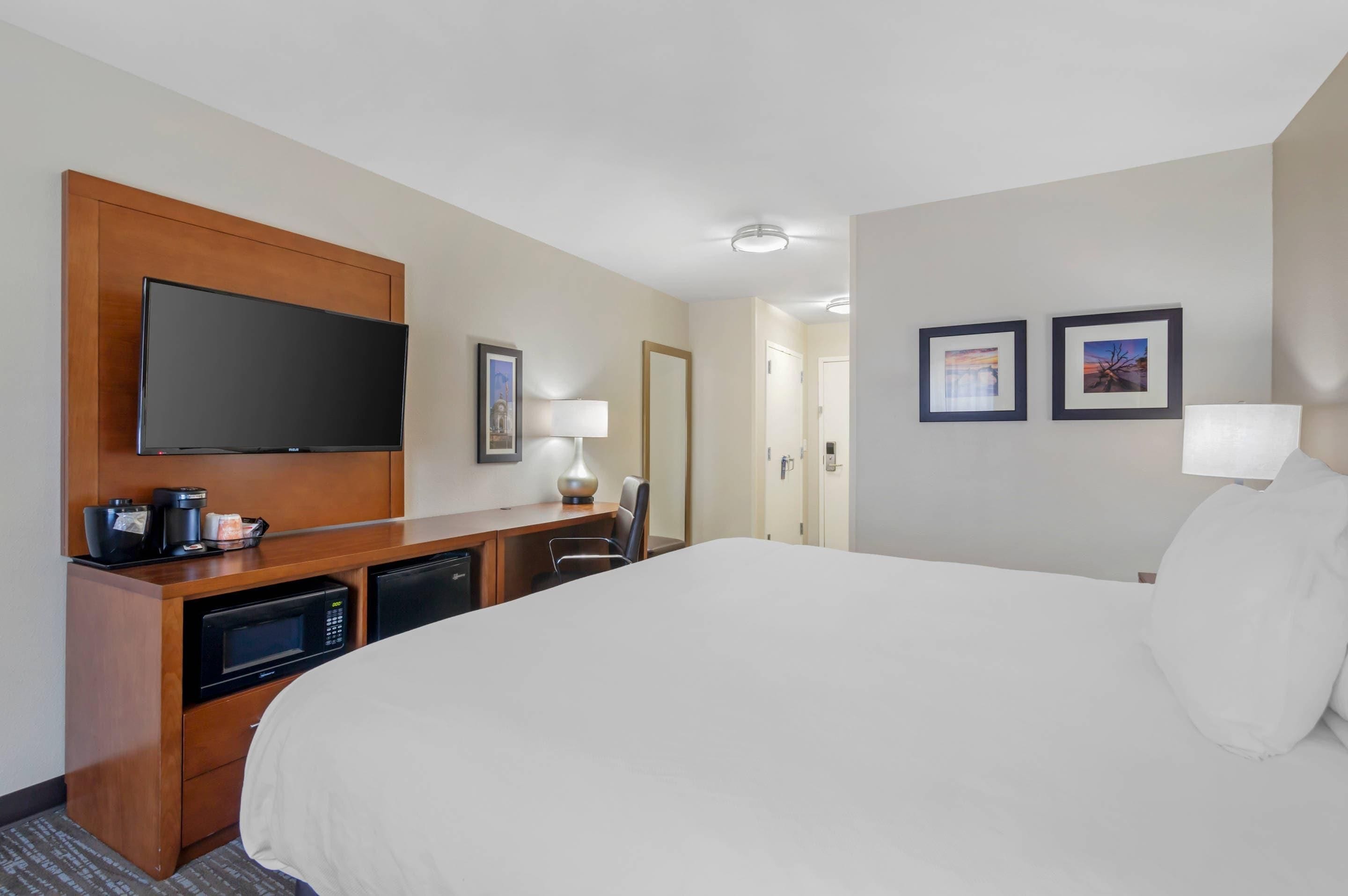 Een hotelkamer met een groot bed, een flatscreen televisie bevestigd aan een houten paneel, een bureau met een koffiezetapparaat, een magnetron en een stoel. Twee ingelijste foto’s hangen aan de muur.