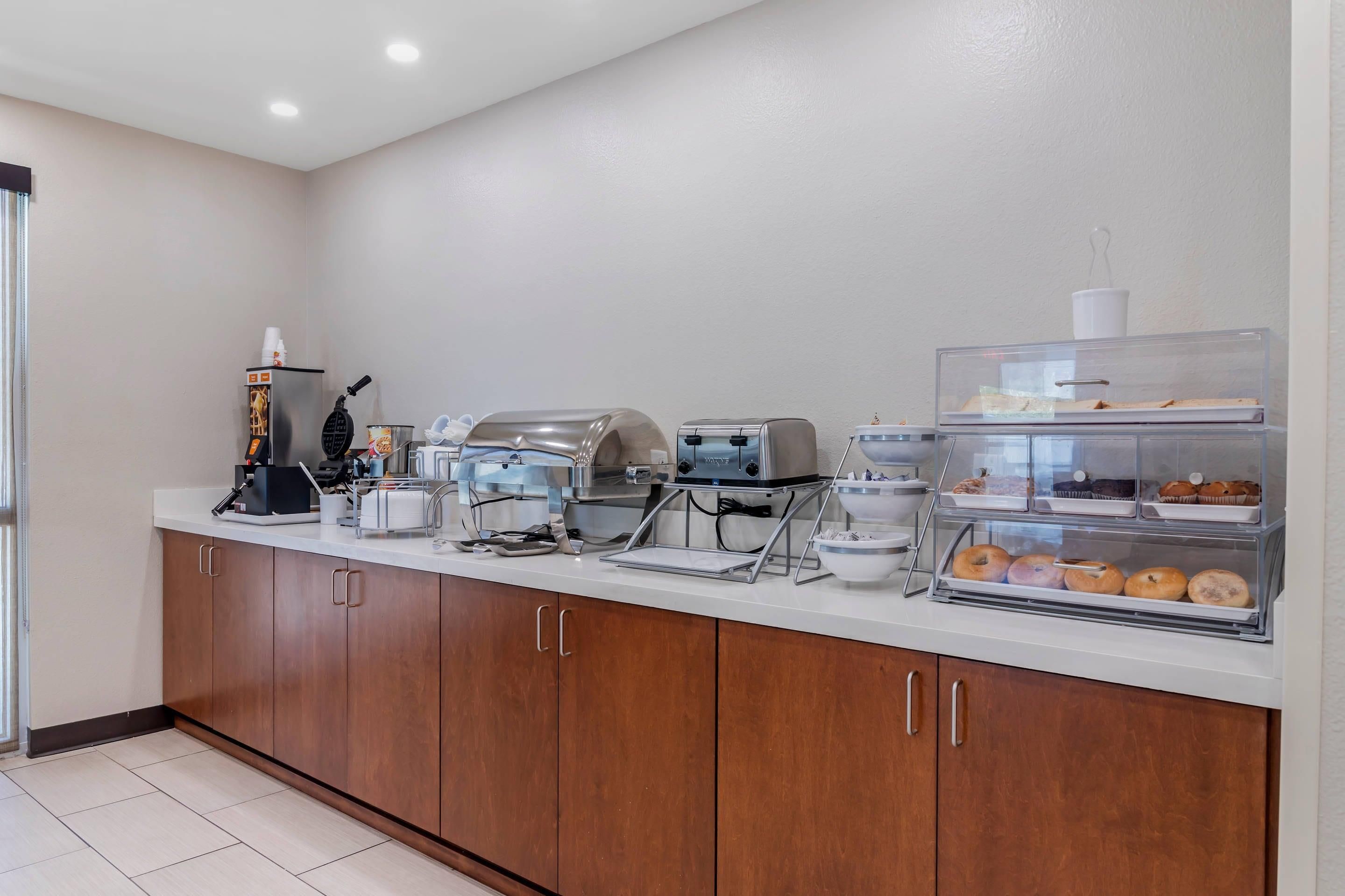 A breakfast bar with a coffee machine, waffle maker, toaster, and a display case with pastries.