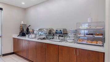A breakfast bar with a coffee machine, waffle maker, toaster, and a display case with pastries.
