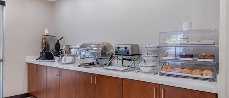 A breakfast bar with a coffee machine, waffle maker, toaster, and a display case with pastries.