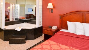 A hotel room with a large bed, a wooden headboard, a nightstand with a lamp, a bathroom with a large mirror, a bathtub, and a red patterned bedspread.