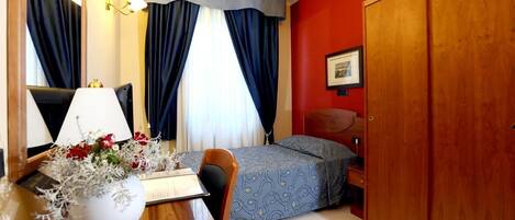 Habitación de hotel con una cama, un escritorio con lámpara, una silla, un armario y un cuadro en la pared.