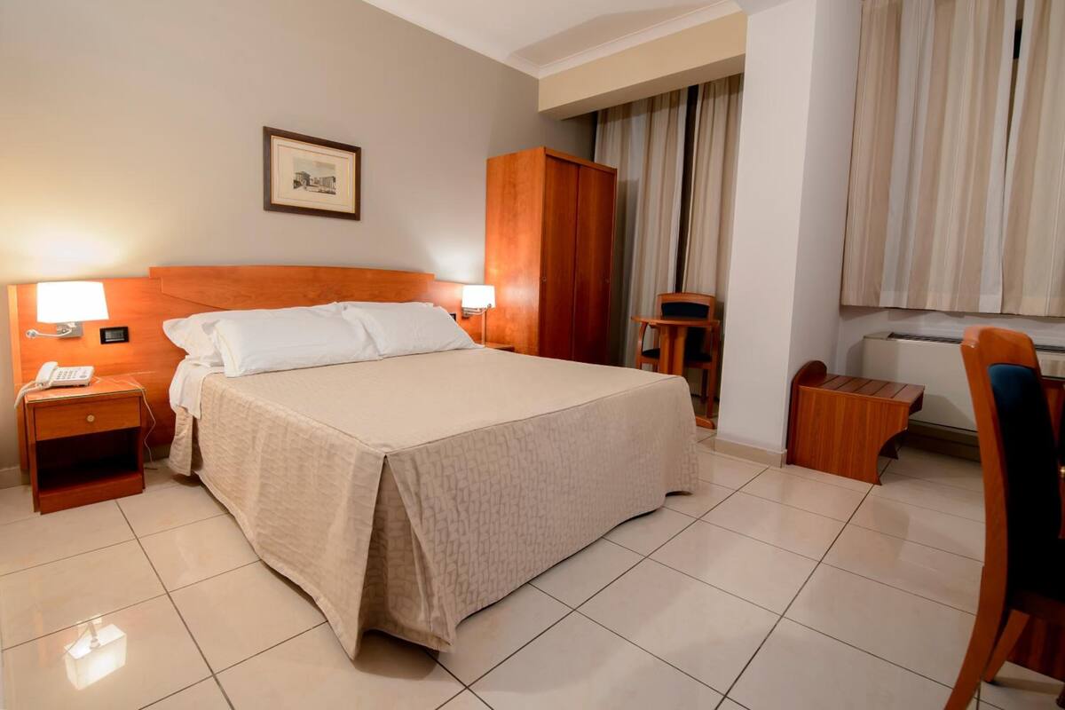 Une chambre d’hôtel avec un grand lit, des lampes de chevet, une armoire en bois, une petite table, une chaise et un tableau encadré au mur.