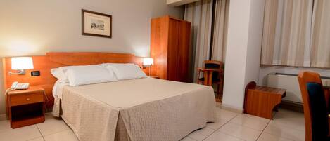 Habitación de hotel con una cama grande, lámparas de noche, un armario de madera, una mesita, una silla e imagen enmarcada en la pared.