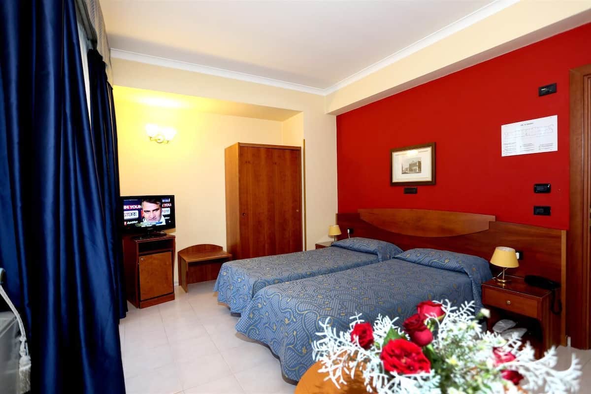 Une chambre d’hôtel avec deux lits, une télévision, une armoire en bois, un bureau avec une lampe et une petite table sur laquelle est posé un vase de fleurs.